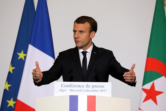 Emmanuel Macron delivers a speech in Algiers, surrounded by French and Algerian flags.