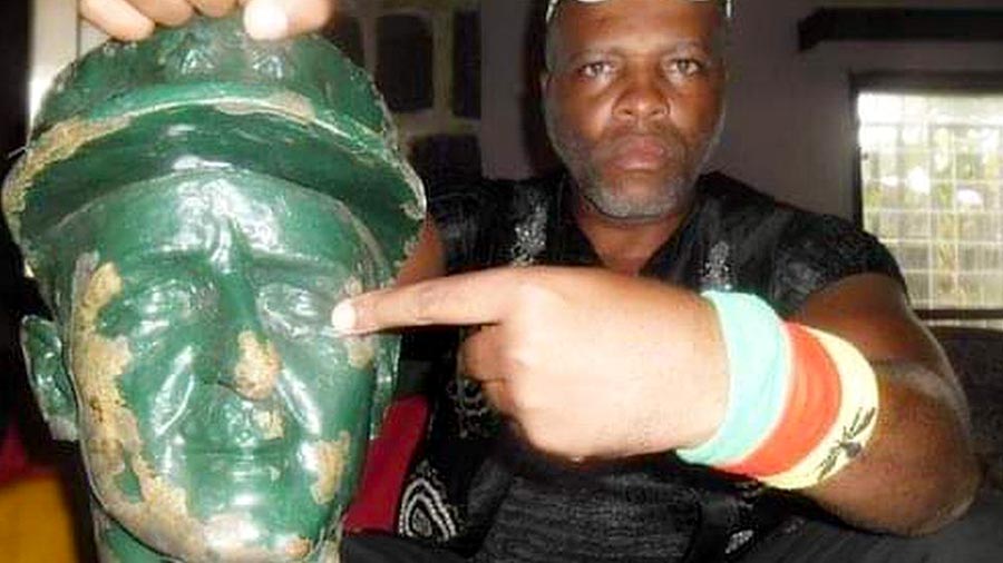 André Blaise Essama proudly holds in his hand the head of a bronze statue, which he points to.