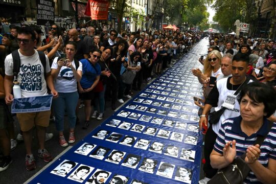 Argentina is at the forefront of universal jurisdiction, partly due to its troubled history of political violence. Photo: In a crowded street, a very long banner is laid out on the ground displaying dozens of black and white portraits.