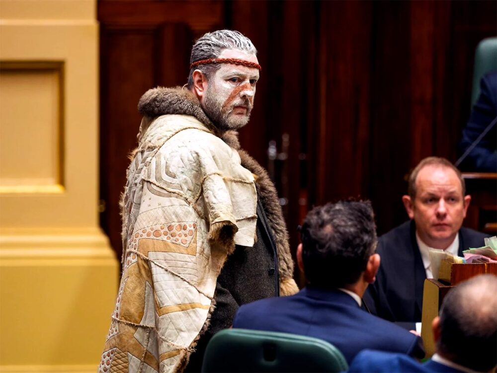 l’Assemblée des Peuples Premiers de Victoria s'exprime devant le Parlement de l’État de Victoria. Photo : Marcus Stewart (co-président de l'Assemblée), portant des habits traditionnels des Peuples Autochtones australien, prononce un discours, entouré de personnalités politiques en costumes plus 'classiques'.