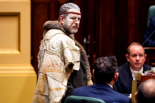 l’Assemblée des Peuples Premiers de Victoria s'exprime devant le Parlement de l’État de Victoria. Photo : Marcus Stewart (co-président de l'Assemblée), portant des habits traditionnels des Peuples Autochtones australien, prononce un discours, entouré de personnalités politiques en costumes plus 