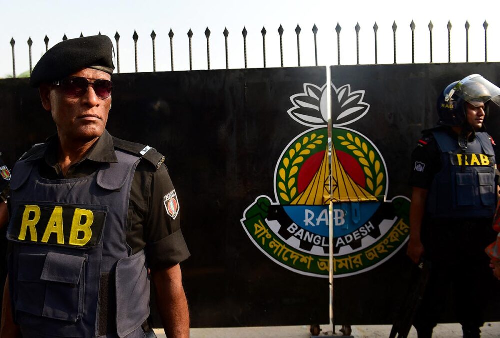 Entrée du camp du Bataillon d'action rapide (RAB) à Dhaka, au Bangladesh. Photo : deux gardes en uniforme portant les lettres « RAB » montent la garde à l'entrée du camp, devant un grand portail en fer noir arborant le blason de l'unité.