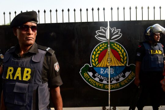 Entrée du camp du Bataillon d'action rapide (RAB) à Dhaka, au Bangladesh. Photo : deux gardes en uniforme portant les lettres « RAB » montent la garde à l'entrée du camp, devant un grand portail en fer noir arborant le blason de l'unité.