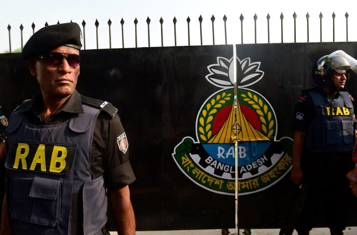 Entrée du camp du Bataillon d'action rapide (RAB) à Dhaka, au Bangladesh. Photo : deux gardes en uniforme portant les lettres « RAB » montent la garde à l'entrée du camp, devant un grand portail en fer noir arborant le blason de l'unité.
