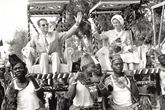 Belgian Colonial Past - Black and white photo in which King Baudouin and Queen Fabiola of Belgium are sitting on armchairs carried by Congolese in traditional dress.
