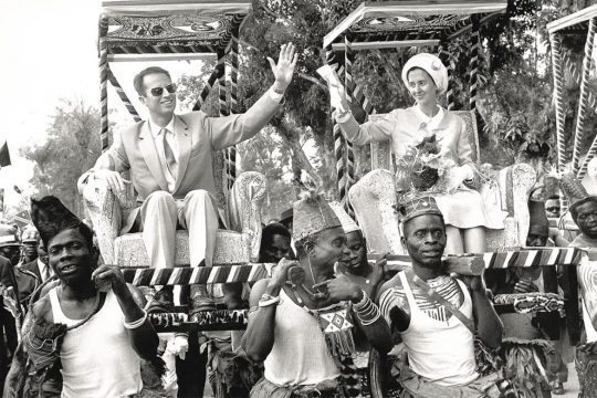 Passé colonial belge - Photo en noir et blanc dans laquelle le Roi Baudouin et la Reine Fabiola de Belgique sont assis sur des fauteuils portés par des congolais en tenues traditionnelles.