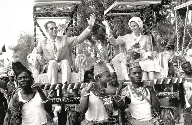 Passé colonial belge - Photo en noir et blanc dans laquelle le Roi Baudouin et la Reine Fabiola de Belgique sont assis sur des fauteuils portés par des congolais en tenues traditionnelles.