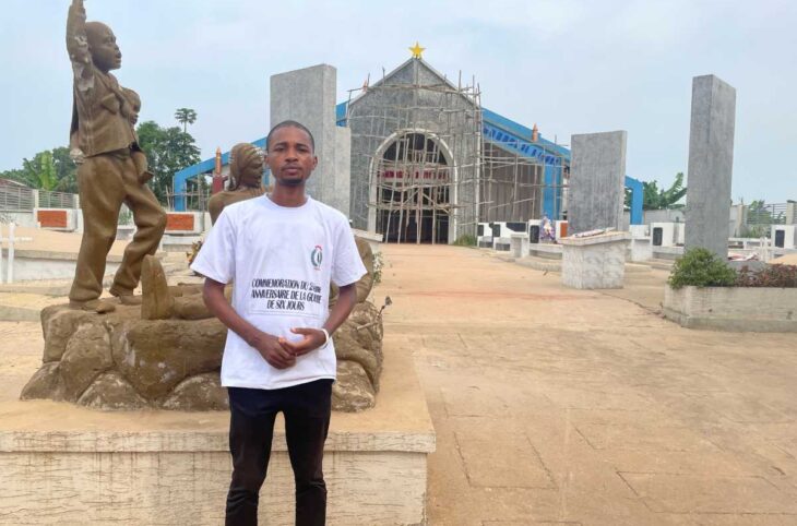 Blaise Monduka, en t-shirt blanc de commémoration de la guerre des six jours, debout devant le Mémorial aux victimes à Kisangani, à côté d'une statue au poing levé.