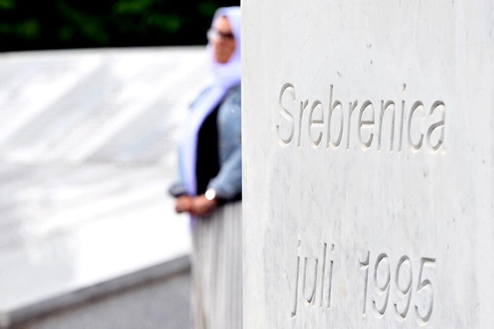 Srebrenica 30 years on: who was convicted of the genocide? Photo: in the foreground, the inscription ‘Srebrenica - Juli 1995’ and in the background, a veiled woman meditates in front of the memorial displaying the names of the victims.