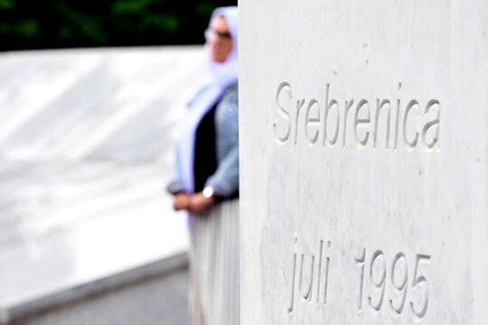Srebrenica 30 years on: who was convicted of the genocide? Photo: in the foreground, the inscription ‘Srebrenica - Juli 1995’ and in the background, a veiled woman meditates in front of the memorial displaying the names of the victims.