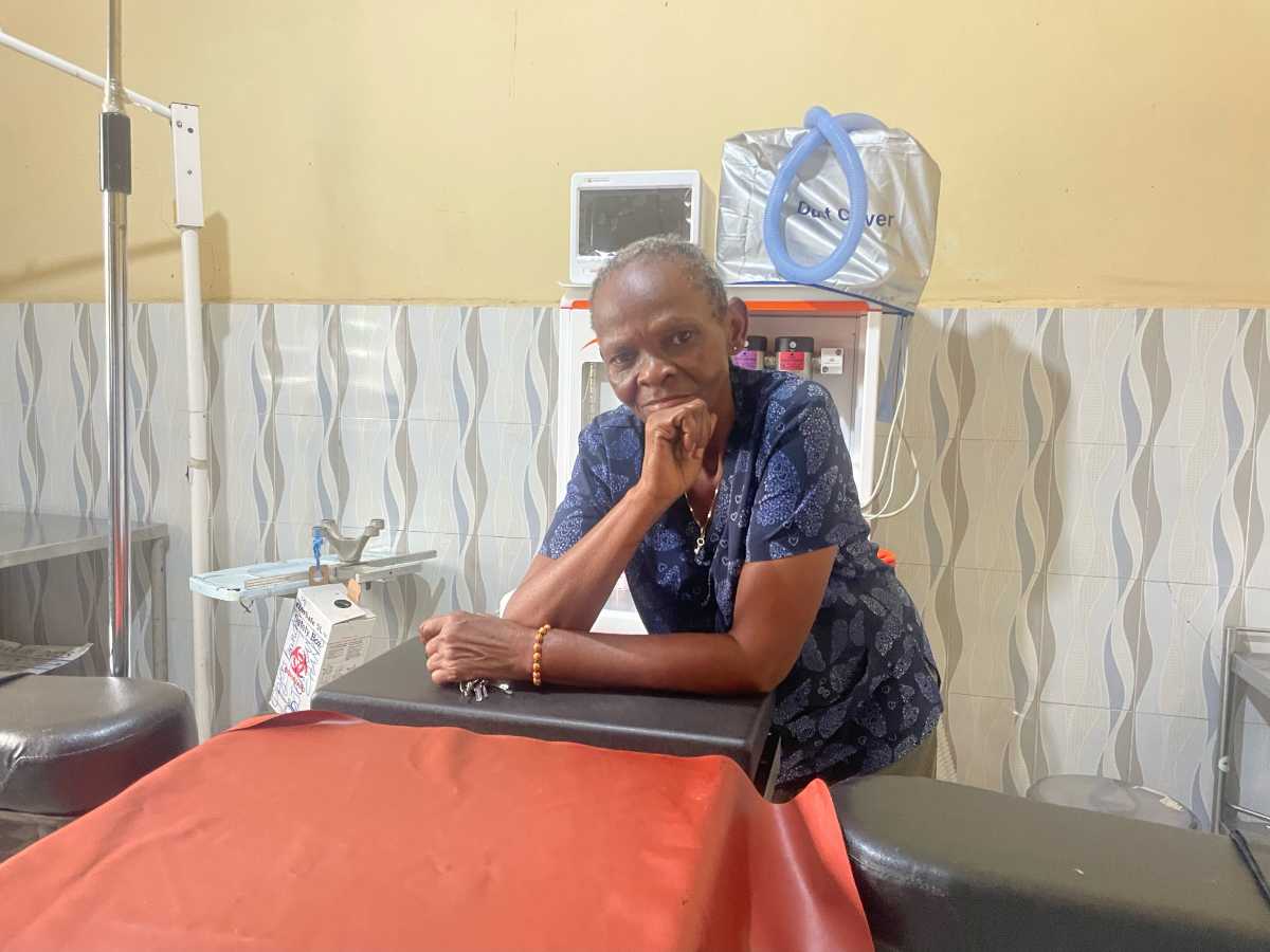 Charly Yenga, femme âgée en robe bleue à motifs, appuyée sur une table d'examen dans une salle médicale, la tête posée sur la main ; en arrière-plan, un moniteur et du matériel de soins.