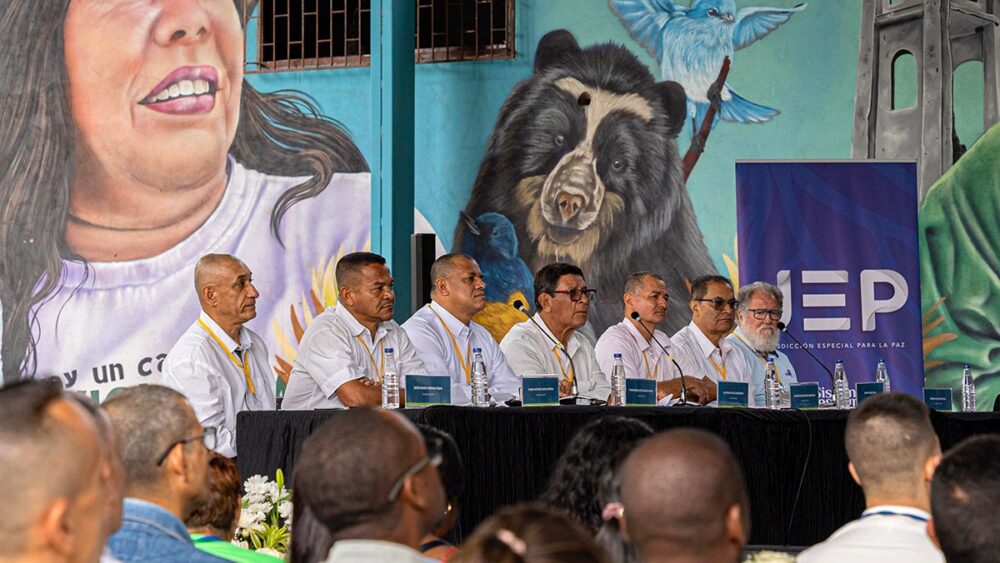 En Colombie, des victimes d'enlèvements se retrouvent face aux commandants FARC responsables de ces crimes. Photo : 7 anciens commandants de la guérilla sont assis, alignés devant un bureau et des micros, en chemises blanches et regardent devant eux. Derrière eux, une fresque murale représentant des hommes et des animaux.