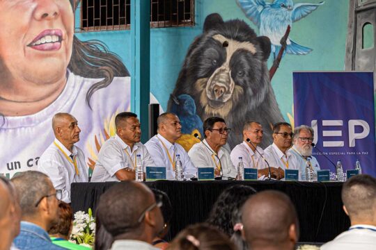 En Colombie, des victimes d'enlèvements se retrouvent face aux commandants FARC responsables de ces crimes. Photo : 7 anciens commandants de la guérilla sont assis, alignés devant un bureau et des micros, en chemises blanches et regardent devant eux. Derrière eux, une fresque murale représentant des hommes et des animaux.