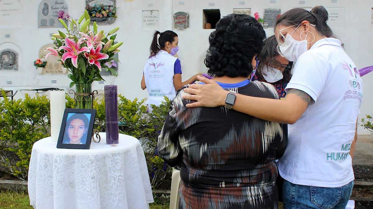Dans un cimetière, une personne de l'équipe UBPD enlace Arnobia Gutiérrez au moment de l'exhumation de la tombe de sa fille (en photo sur une table avec des fleurs et des bougies).