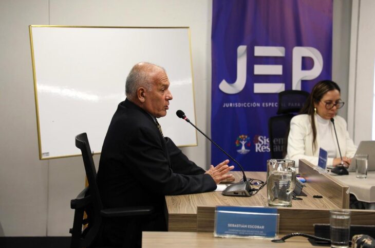 La Juridiction spéciale pour la paix de Colombie (JEP : le mécanisme pénal de la justice transitionnelle) a condamné le colonel retraité Hernán Mejía a 20 ans de prison. Photo : Mejía s'exprime devant un micro. En arrière-plan un panneau affichage le logo de la JEP.