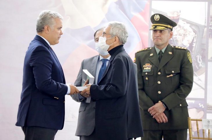 President Iván Duque stands in front of Truth Commission chairman father Francisco de Roux. They exchange a document (defence sector's truth report) under the gaze of a military man.