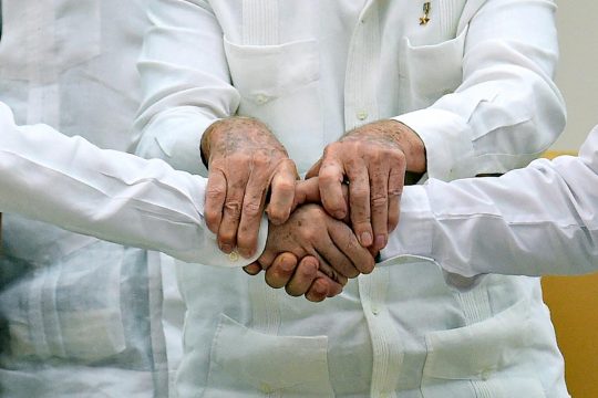 Deux hommes se serrent la main avec l'aide d'un troisième, tous vêtus de chemises blanches (symbole de paix en Colombie)