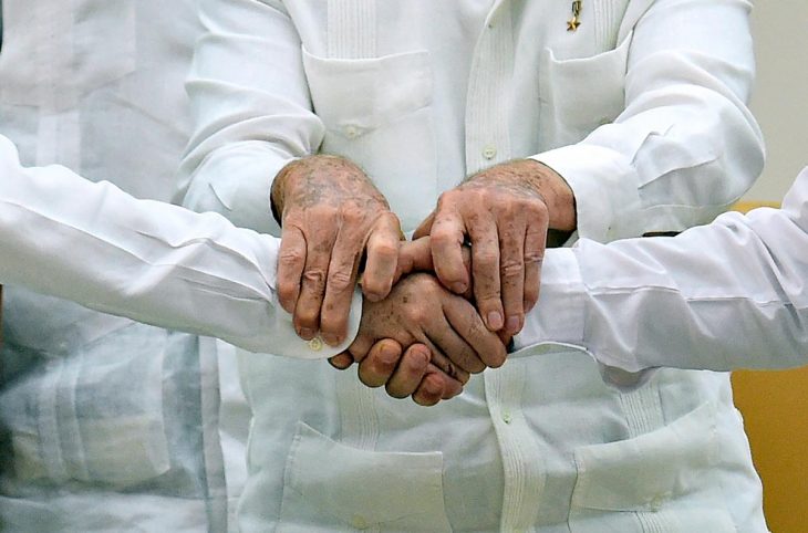 Deux hommes se serrent la main avec l'aide d'un troisième, tous vêtus de chemises blanches (symbole de paix en Colombie)
