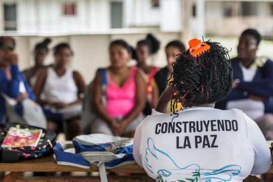 A woman (from behind) wearing a "construyendo la paz" t-shirt is facing several people who seem to be listening to her