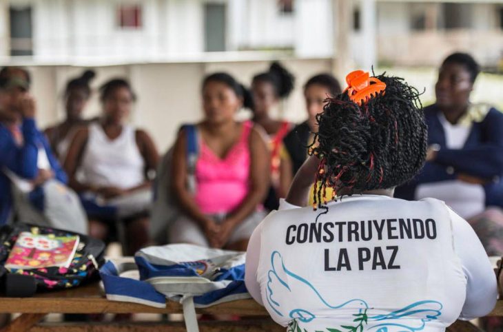 Une femme (de dos) portant un t-shirt "construyendo la paz" fait face à plusieurs personnes qui semblent l'écouter