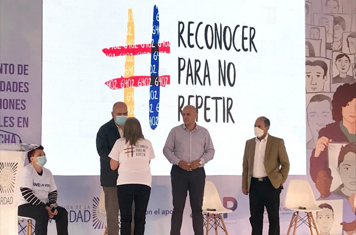 During an event of the Truth Commission of Colombia, three ex-Colombian officers, standing on a platform, meet a woman (she is shaking hands with one of them). In the background, a large screen shows the words "reconocer para no repetir" in Spanish.