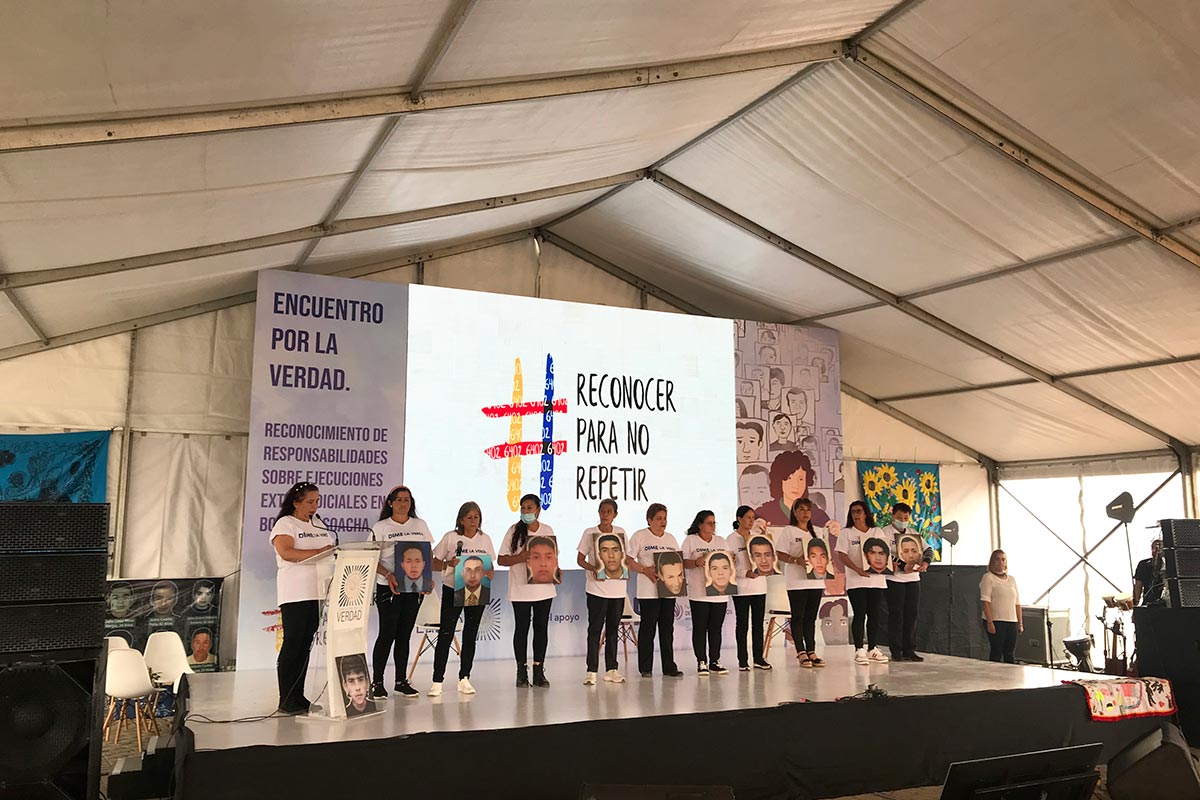 11 mothers of victims of the "false positives" scandal are standing on a platform (under a marquee) holding pictures of their sons murdered by the military. Behind them, a large screen reads, in Spanish, "reconocer para no repetir" and "encuentro por la verdad".