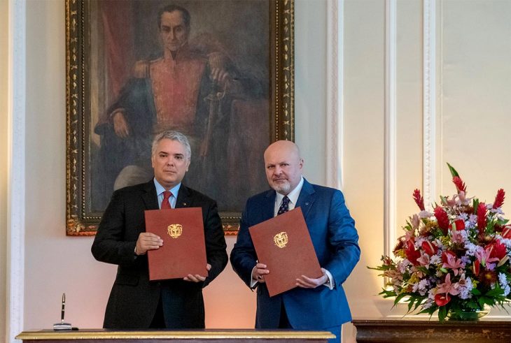 Iván Duque and Karim Khan pose with a document in hand. A painting of Simon Bolivar is displayed behind them.