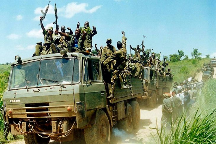 Ugandan soldiers in military transport trucks, waving fists and weapons