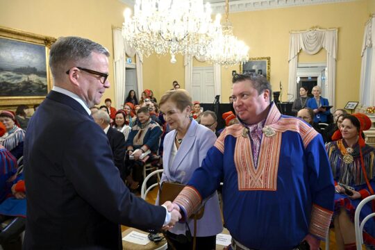 En Finlande, le premier ministre Petteri Orpo et Tuomas Aslak Juuso (du parlement Sami) se sont rencontrés pour la remise du rapport de la Commission vérité et réconciliation. Photo : Orpo et Juuso se serrent la main dans une salle de réception comble. Juuso porte les vêtements traditionnels samis.
