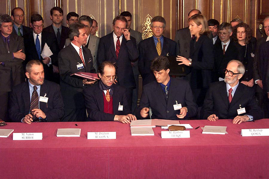 In 1999, Albanian negotiators from Kosovo (including Hashim Thaçi) travelled to France (to Rambouillet, near Paris) to sign a peace agreement. Photo: a group of people in suits stand behind a table where Thaçi, Rugova, Surroi and Qosja are preparing to sign the agreement.