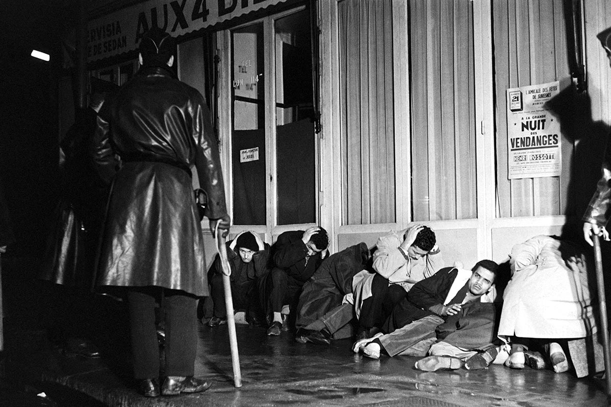 La police surveille, bâton à la main, des manifestants algériens assis sur le sol, mains sur la tête, devant une boutique parisienne en 1961.