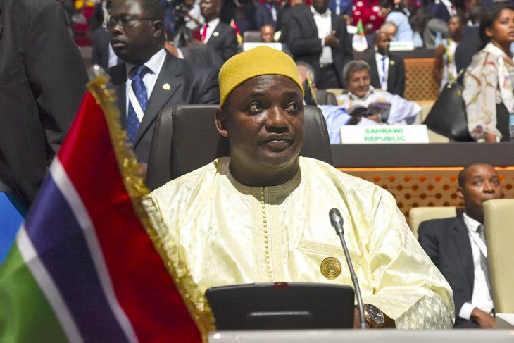 Gambian President Adama Barrow sits in a chair at an international diplomatic event. In the foreground: a small flag of Gambia.
