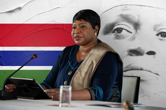 Fatou Bensouda sits in front of a microphone. In the background, a portrait of Yahya Jammeh and the Gambian flag.