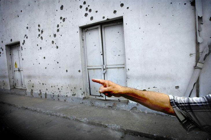 Quelqu'un montre du doigt quelque chose. En arrière-plan, des impacts d'armes dans un mur.