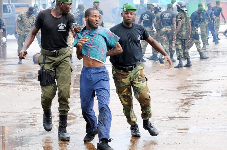 Dans une rue de Conakry en Guinée, près du stade, deux hommes en uniforme militaire ont arrêté un civil. En arrière-plan d'autres hommes en uniforme.
