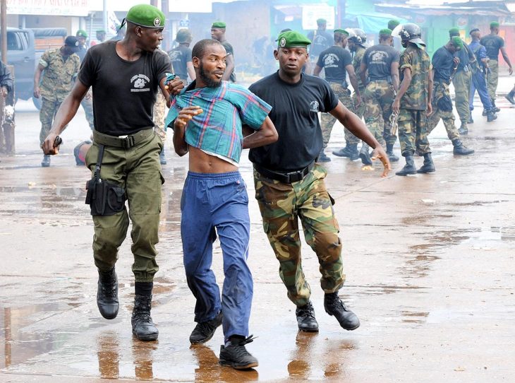 Dans une rue de Conakry en Guinée, près du stade, deux hommes en uniforme militaire ont arrêté un civil. En arrière-plan d'autres hommes en uniforme.