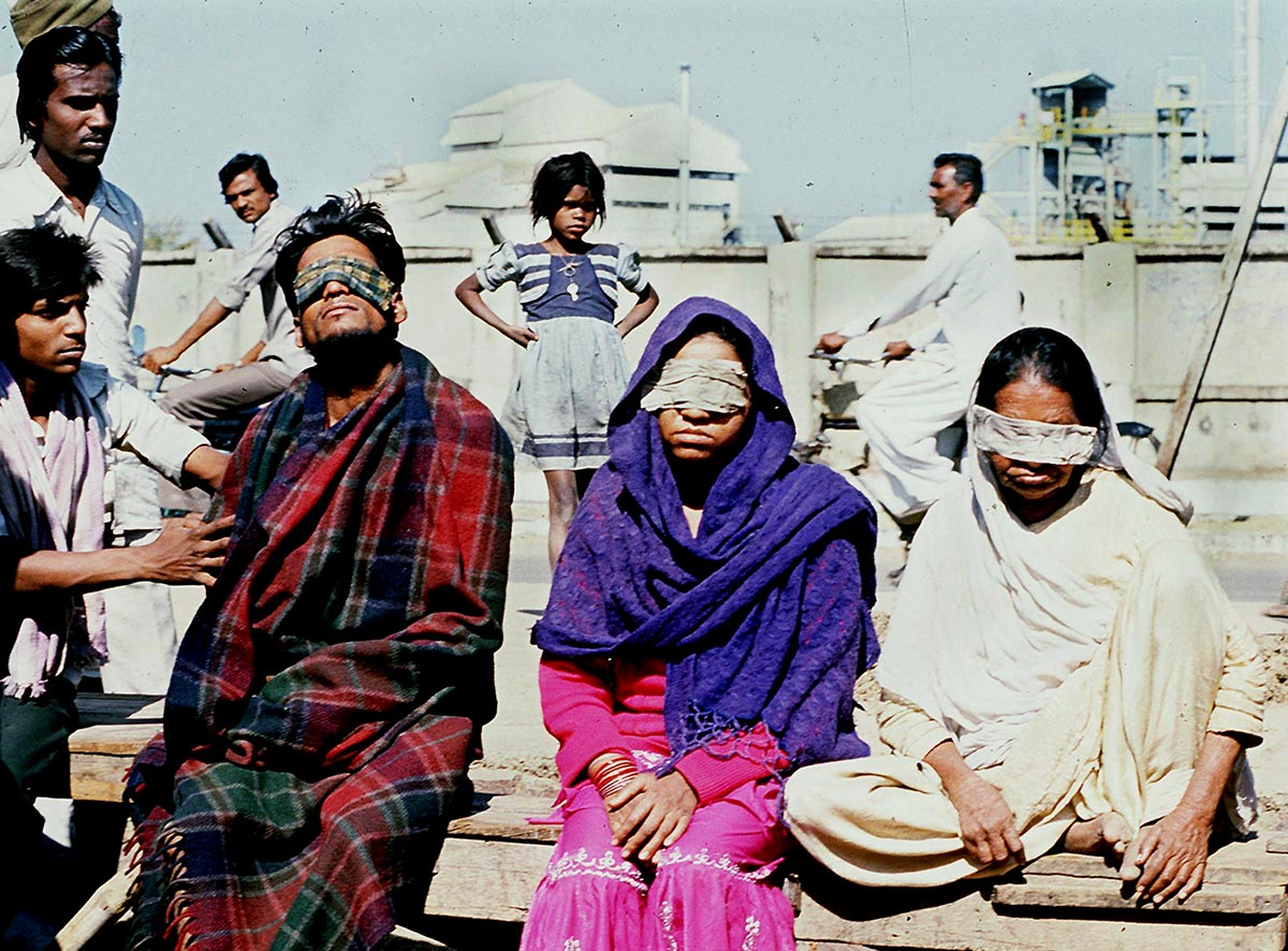 Hommes et femmes portant des lambeaux de tissus sur les yeux. En arrière-plan une grande usine, celle de Bophal en Inde.