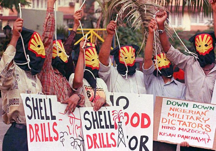 Masked protesters (Shell logo) raise their fists and hold up "Shell drills" signs.