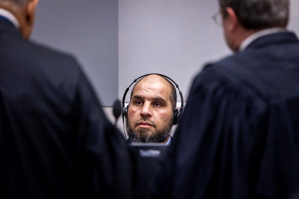 Le Libyen Khaled Mohamed Ali El Hisri, accusé notamment de crimes contre l’humanité contre des migrants à la prison de Mitiga à Tripoli, dialogue avec ses avocats lors de l’audience dite de “confirmation des charges”, la semaine dernière, à La Haye. Photo : © ICC-CPI La Cour pénale internationale (CPI) enquête en Libye depuis 15 ans sur les crimes commis contre les migrants. Photo : Khaled Mohamed Ali El Hishri (assis avec un casque sur les oreilles), lors de son procès à La Haye, écoute deux avocats, de dos.
