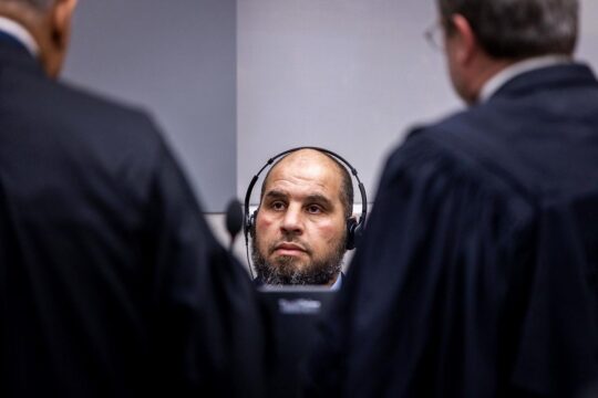 La Cour pénale internationale (CPI) enquête en Libye depuis 15 ans sur les crimes commis contre les migrants. Photo : Khaled Mohamed Ali El Hishri (assis avec un casque sur les oreilles), lors de son procès à La Haye, écoute deux avocats, de dos.