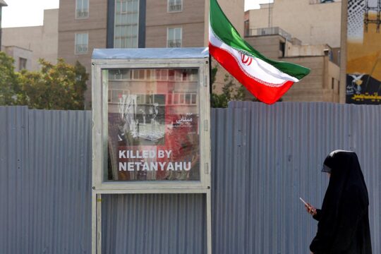 Aggression by Israel and the United States in Iran - Photo: portrait of a victim of an Israeli attack in a street in Tehran (Iranian flag and veiled woman walking while looking at her smartphone).