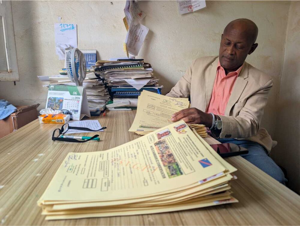 Jean Kalegamire Bienda, vêtu d'une veste beige et d'une chemise rose, assis à son bureau et examinant une pile de dossiers de victimes ; derrière lui, des piles de documents et un calendrier ouvert à juin 2026.