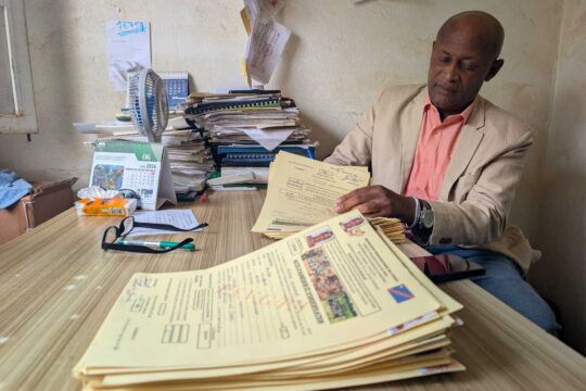 Jean Kalegamire Bienda, vêtu d'une veste beige et d'une chemise rose, assis à son bureau et examinant une pile de dossiers de victimes ; derrière lui, des piles de documents et un calendrier ouvert à juin 2026.