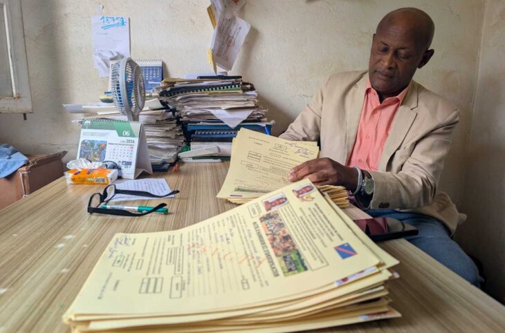 Jean Kalegamire Bienda, vêtu d'une veste beige et d'une chemise rose, assis à son bureau et examinant une pile de dossiers de victimes ; derrière lui, des piles de documents et un calendrier ouvert à juin 2026.