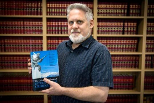 Kevin Jon Heller holds a book in his hands (he is the co-author): "The Oxford handbook of international criminal law". In the background: a library.