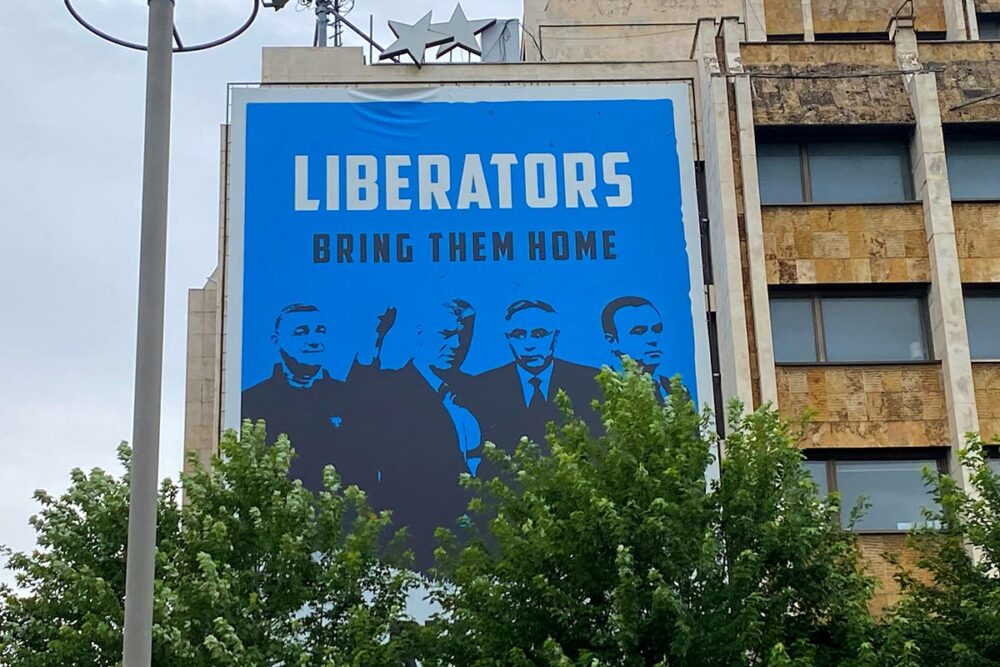 Une affiche appelle au retour au pays des « libérateurs », qui attendent actuellement leur jugement devant les Chambres spécialisées du Kosovo (KSC), sur le Grand Pristina Hotel, dans le centre de la capitale du Kosovo. Photo prise le 8 juillet 2026. Photo : © Serbeze Haxhiaj Les Chambres spécialisées du Kosovo (KSC) : un tribunal spécial (basés à La Haye) pour les crimes de guerre et crimes contre l'humanité commis par les forces kosovares durant la guerre contre la Serbie, dont l'objet et le fonctionnement sont controversés. Photo : une grande affiche bleue est accrochée à un immeuble. Il y est écrit : 'Liberators. Bring them home.' On reconnait le visage d'Hashim Thaçi et de ses 3 co-accusés devant les KSC.