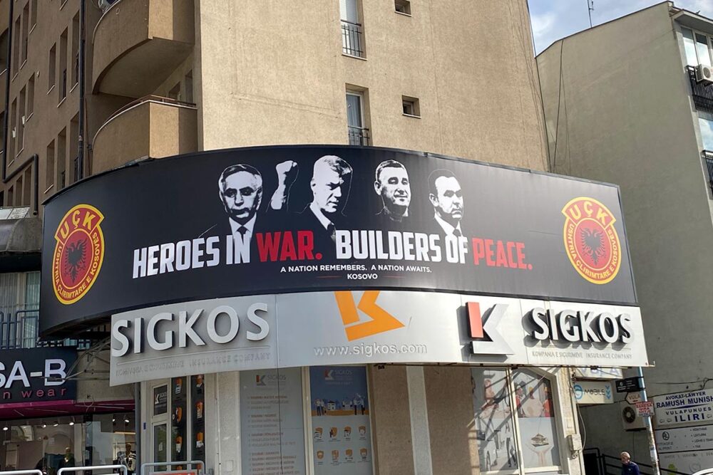 Banderole rendant hommage aux « héros de guerre », « artisans de la paix » que « toute une nation attend », photographiée le 11 octobre 2026 dans le centre de Pristina, capitale du Kosovo. Les quatre ex-membres de l’Armée de libération ont été reconnus coupables de crimes de guerre le 16 septembre dernier par les Chambres spécialisées, basées à La Haye. Photo : © Serbeze Haxhiaj Au Kosovo, 4 ex-membres de l'Armée de libération du Kosovo (UCK), condamnés à La Haye pour des crimes de guerre, sont célébrés dans une bannière affichée en pleine rue à Pristina. Photo : une bannière montrant les 4 visages des « héros de guerre » et « artisans de la paix » est accrochée sur la façade d'un immeuble.
