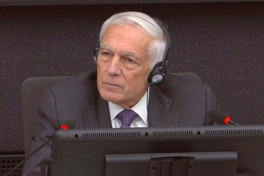 Wesley Clark, un ancien général américain de l'OTAN, témoigne en faveur d'Hashim Thaçi lors de son procès devant les Chambres spécialisées du Kosovo (KSC). Photo : Clark porte un casque sur les oreilles, assis face à des écrans et des micros.