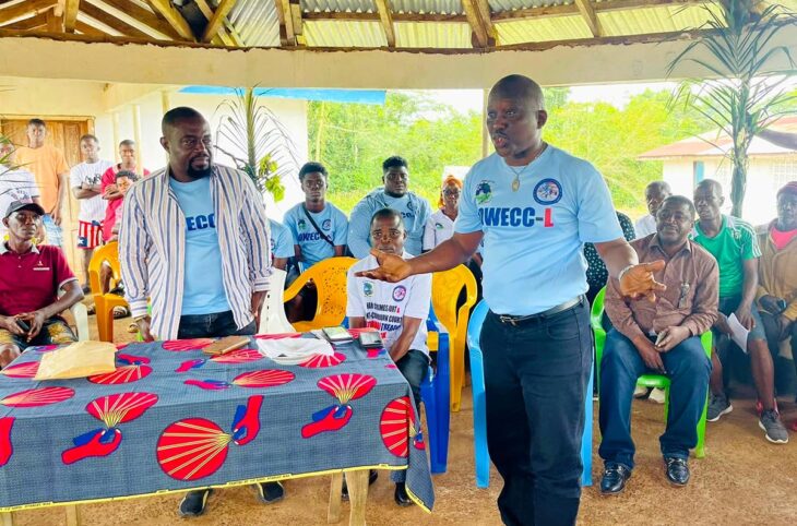 séance de sensibilisation du Bureau de la Cour pour les crimes de guerre et les crimes économiques du Libéria (OWECC-L). Photo : des hommes, dont la plupart portent un t-shirt affichant le logo de l'OWECC-L, sont rassemblés sous un abris en bois et tôle pour écouter un homme.
