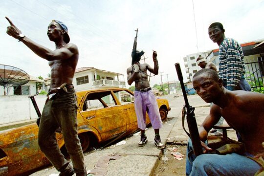 Au Liberia, des combattants du Front national patriotique du Liberia (NPFL), lourdement armés, sont rassemblés autour d'une épave de voiture. L'un d'entre eux semble s'adresser de façon virulente à une autre personne plus loin.